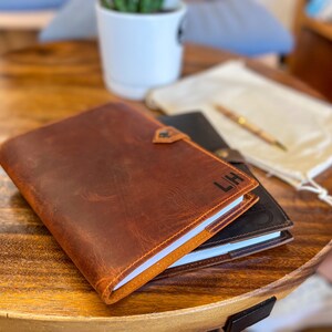 May include: A brown leather journal with the initials "LH" embossed on the cover. The journal is closed and lying on a wooden table.