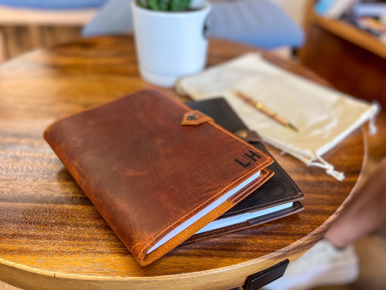 May include: A brown leather journal with the initials "LH" embossed on the cover. The journal is closed and lying on a wooden table.