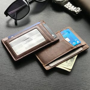 May include: Two brown leather wallets, one open and one closed, are displayed on a dark surface. The open wallet reveals credit cards and a twenty-dollar bill. The closed wallet has a clear ID window and a card visible. Sunglasses and a watch are in the background.