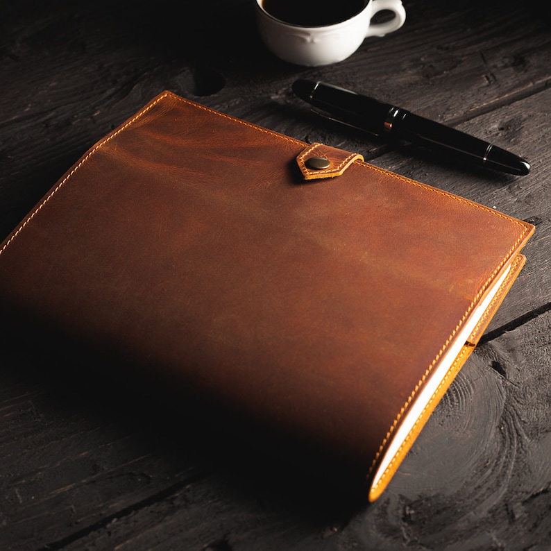 May include: A brown leather journal with a brass button closure. The journal is lying on a dark wooden surface with a pen and a coffee cup.