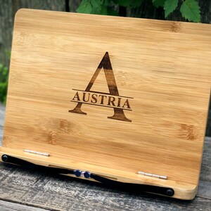 May include: A bamboo book stand with a black strap and metal hinges. The stand is engraved with the letter "A" and the word "AUSTRIA".
