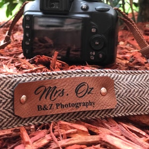 May include: A brown and white camera strap with a brown leather patch that says "Mrs. Oz B&Z Photography" in black lettering.