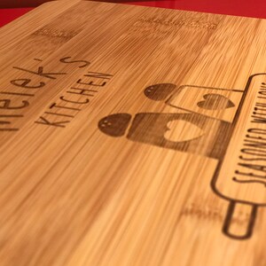 May include: A wooden cutting board with the text "Melek's Kitchen" and "Seasoned with Love" engraved on it. The board features a rolling pin and three salt and pepper shakers with hearts.