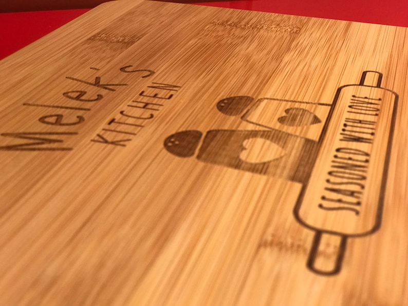 May include: A wooden cutting board with the text "Melek's Kitchen" and "Seasoned with Love" engraved on it. The board features a rolling pin and three salt and pepper shakers with hearts.
