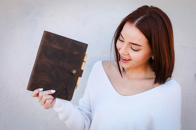 May include: A brown leather journal with a pen holder and a brass button closure. The journal has a personalised monogram on the front.