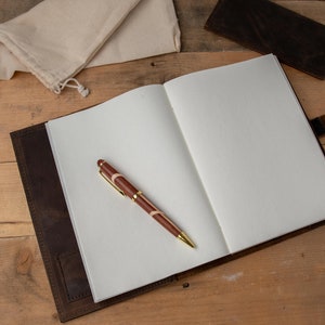 May include: A brown leather journal with a pen lying on the open pages. The journal has a flap closure and a strap with a metal button.