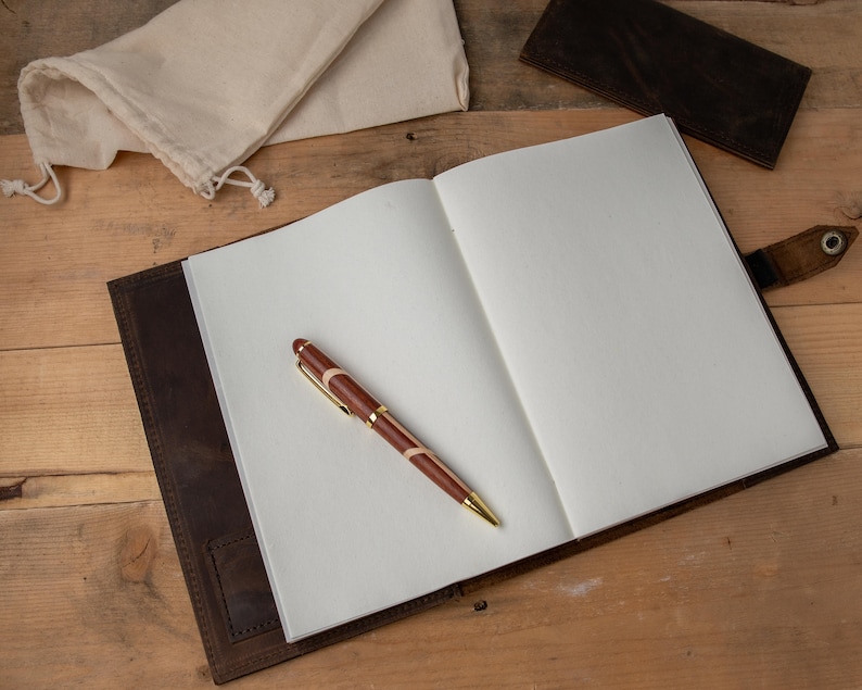 May include: A brown leather journal with a pen lying on the open pages. The journal has a flap closure and a strap with a metal button.
