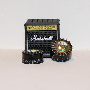 May include: A miniature black Marshall amplifier with gold accents and the brand name in white. Two black knobs with gold and iridescent details are in front of the amp. The knobs have white number markings.