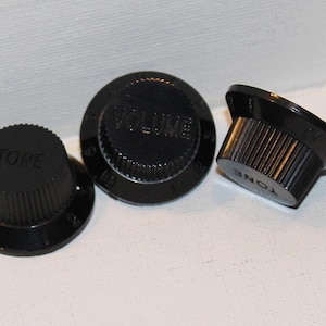 May include: Three black guitar control knobs. One knob reads "TONE", another reads "VOLUME", and the third knob is angled to show the side. The knobs have a ridged edge for grip and are made of a glossy black plastic.