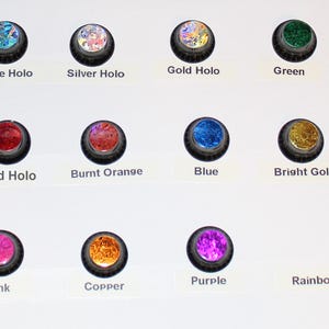 May include: A collection of twelve round, decorative buttons in various colors and finishes. The buttons include Blue Holo, Silver Holo, Gold Holo, Green, Red Holo, Burnt Orange, Blue, Bright Gold, Pink, Copper, Purple, and Rainbow. Each button has a black rim and a shiny, textured center.