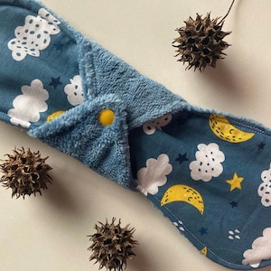 Puede incluir: Una compresa menstrual de tela reutilizable azul con un patrón de luna y nubes. La compresa tiene un cierre a presión y está hecha de tela suave y absorbente.