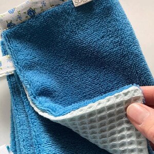 May include: A blue and white kitchen towel with a waffle weave pattern on one side and a soft terry cloth on the other. The towel has a floral print border and a small label that says "Strikset".