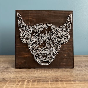 May include: A string art piece featuring a Highland cow head silhouette. The design is crafted with white string on a dark brown stained wooden square. The artwork is displayed on a wooden surface against a blue wall.