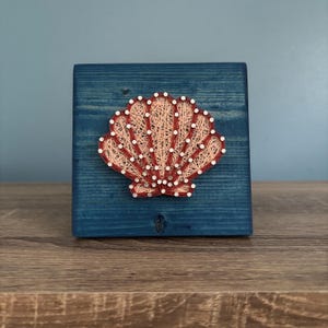 May include: A handcrafted string art piece featuring a seashell design. The seashell is made with peach and red thread, mounted on a square, blue-painted wooden block. The artwork is a decorative item with a nautical theme.