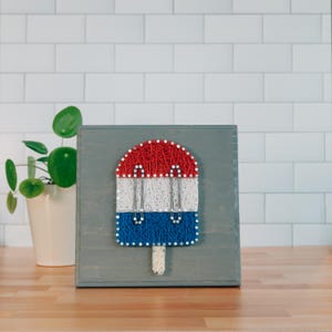 May include: A wooden string art piece depicting a popsicle in red, white, and blue. The popsicle is mounted on a gray wooden block. A white potted plant is visible in the background. The piece is a patriotic decoration.