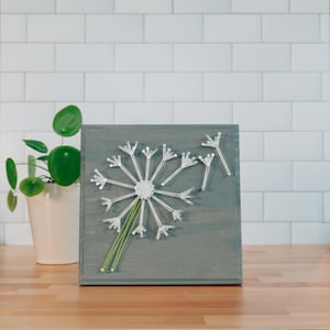 Puede incluir: Una pieza de arte de hilo de diente de león enmarcada en madera gris. El diente de león está hecho de hilo blanco y tallos de plástico transparente. El hilo verde forma el tallo. La obra de arte se exhibe sobre una superficie de madera.