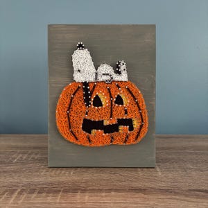 Può includere: Un'opera d'arte con filo raffigurante una zucca arancione intagliata con una faccia sorridente e una figura bianca di Snoopy in cima. L'opera è montata su una tavola di legno grigia, perfetta per Halloween.