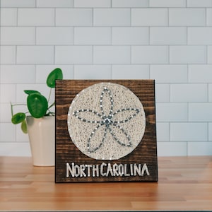May include: A wooden sign featuring a sand dollar design made with string art. The sand dollar is centered on a dark brown square, with the words "NORTH CAROLINA" in white letters at the bottom. The sign is displayed on a wooden surface.