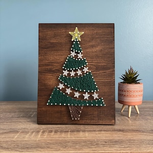 May include: A handcrafted Christmas tree string art piece on a stained wooden board. The tree is made with green string, decorated with white star accents, and topped with a gold star. A festive holiday decoration.