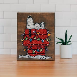 Può includere: Un'opera d'arte in filo che raffigura Snoopy, l'amato beagle dei Peanuts, che dorme sulla cima della sua cuccia rossa. La cuccia è decorata con luci colorate in filo, creando una scena natalizia festosa.