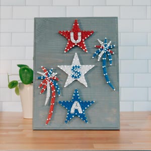 May include: Wooden sign featuring five star-shaped string art designs in red, white, and blue. The stars are arranged on a gray wooden background, with the letters "U" and "A" visible. The sign is a patriotic decoration.