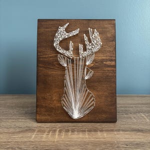 May include: A wooden string art piece featuring a deer head silhouette. The deer is crafted with white string against a dark brown stained wooden background. The artwork is displayed on a wooden surface with a blue wall in the background.