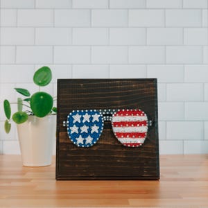 May include: A dark brown wooden box featuring a string art design of sunglasses. The left lens is blue with white stars, and the right lens is red and white striped, resembling the American flag. The sunglasses are attached with silver hardware.