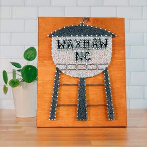 May include: A wooden sign featuring a string art design of a water tower. The water tower is made with white and gray string, with the words "WAXHAW NC" spelled out in black letters. The sign is mounted on a stained wooden board.