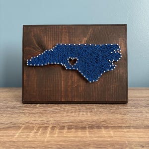 May include: A wooden wall art piece featuring a string art design of the North Carolina state outline. The state is filled with blue string and has a heart-shaped cutout. The wooden frame is stained a dark brown color.