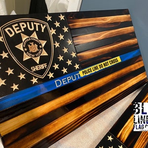 May include: A wooden American flag with a blue line and a sheriff's badge. The badge is gold and has the words "Suffolk County" and "Sheriff" on it. The flag is burned and has a yellow "Police Line Do Not Cross" tape across it. The flag is made by Blue Line Flag Co.