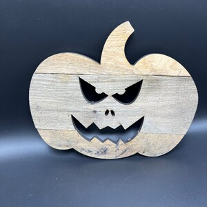 May include: A wooden pumpkin with a carved scary face. The pumpkin is painted in a light brown color and has a black face with a wide grin and two eyes.