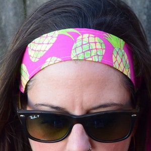 May include: A pink headband with a pineapple print. The headband is worn by a person with dark hair and sunglasses.