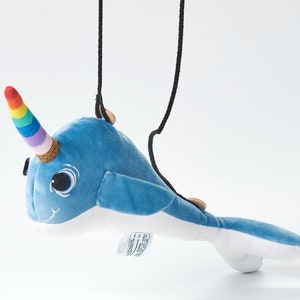May include: A blue plush narwhal with a rainbow horn and black cord. The narwhal has a white belly and large black eyes.