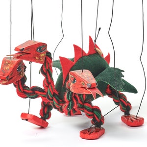 May include: A red, green, and black puppet dragon with three heads and a green felt body. The dragon is made of wood and fabric and has strings attached to its limbs and heads.
