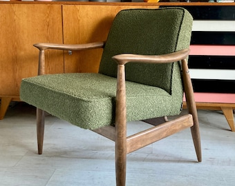 Può includere: Una poltrona imbottita verde con gambe e braccioli in legno. La poltrona è posizionata su un pavimento in legno davanti a un mobile in legno.