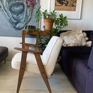 May include: A white armchair with wooden legs and arms. The chair is upholstered in a fluffy white fabric. The chair is in a living room setting with a couch and a plant in the background.