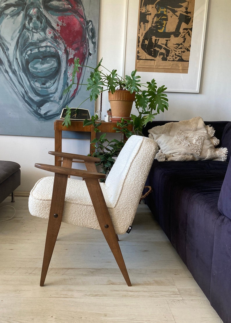 May include: A white armchair with wooden legs and arms. The chair is upholstered in a fluffy white fabric. The chair is in a living room setting with a couch and a plant in the background.