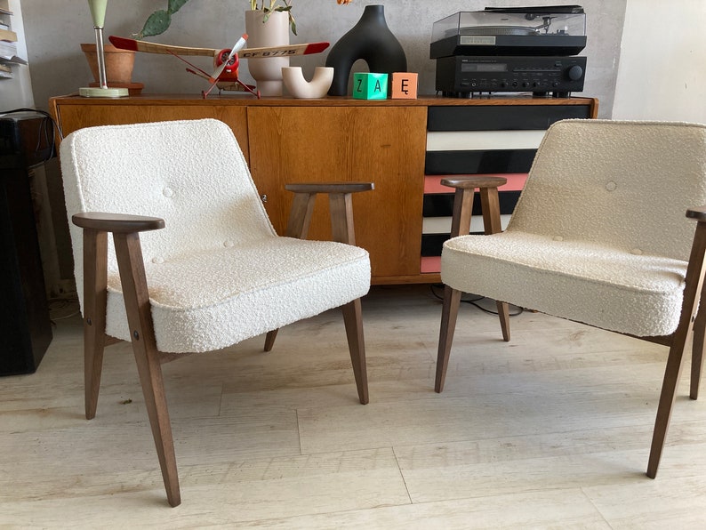 May include: Two white chairs with wooden legs and arms. The chairs have a textured fabric upholstery and are positioned in front of a wooden cabinet with black and white stripes.