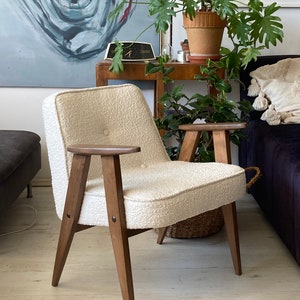 May include: A white armchair with a textured fabric upholstery and wooden legs. The chair has a button tuft on the back and a single button on the armrest.