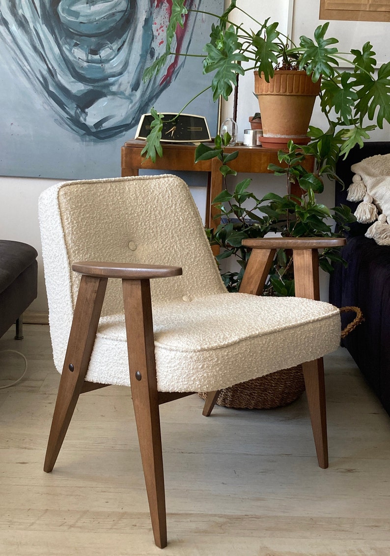 May include: A white armchair with a textured fabric upholstery and wooden legs. The chair has a button detail on the back and wooden armrests.