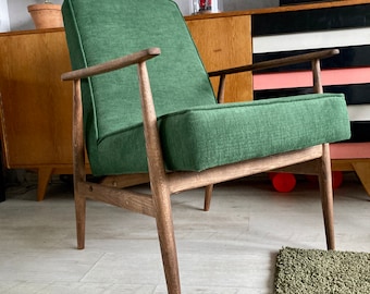 May include: A green upholstered armchair with dark wood legs. The chair is sitting on a light wood floor with a green rug in the foreground.