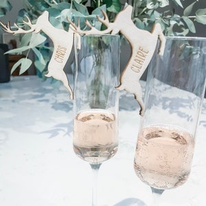 Puede incluir: Dos copas de champán con porta tarjetas de madera en forma de reno. Los renos están grabados con los nombres "Chris" y "Claire".