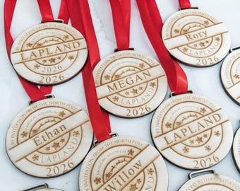 May include: Round wooden ornaments with red ribbons, each engraved with the text BY INVITATION FROM THE NORTH POLE A MAGICAL ADVENTURE, LAPLAND, 2026, and various names including Ethan, Megan, Rory, and Willow, featuring star and snowflake motifs.