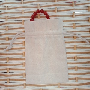 May include: A small, off-white drawstring bag with a red beaded handle. The bag is made of a plain, textured fabric and sits on a woven, light brown surface. The drawstring is a light beige color.