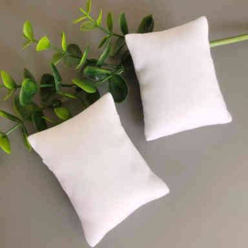 May include: Two white rectangular pillow-shaped cushions, made of soft fabric, are lying on a gray surface.