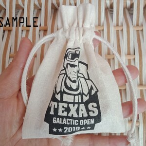 May include: A small, off-white drawstring bag with a black graphic of a futuristic soldier and the text "TEXAS GALACTIC OPEN 2019". The word "SAMPLE" is printed at the top. The bag is held in a hand.