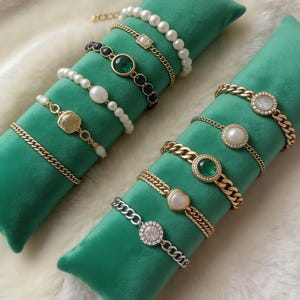 Op de afbeelding: Een assortiment gouden en zilveren armbanden met diverse ontwerpen, waaronder parels, edelstenen en schakels, gepresenteerd op groene fluwelen kussens. De armbanden hebben verschillende versieringen, zoals een hartvormige bedel en een vierkante edelsteen.