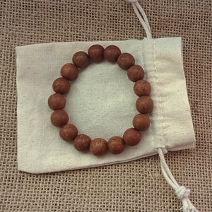 May include: A round wooden bead bracelet with a natural finish rests on a small, off-white drawstring bag. The bracelet is made of smooth, reddish-brown beads. The bag is made of a light-colored fabric and is placed on a textured, tan surface.
