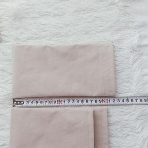 Könnte beinhalten: Beiger Stoffartikel mit einem Metall-Maßband, das Zoll und Zentimeter anzeigt. Der Stoff ist gefaltet und liegt auf einer weißen, flauschigen Oberfläche. Das Maßband ist mit Zahlen markiert.