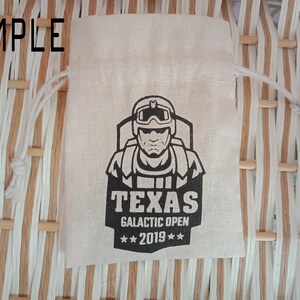 May include: A beige drawstring bag with a black graphic of a person wearing a helmet and goggles. The text "TEXAS GALACTIC OPEN 2019" is printed below the graphic. The word "SAMPLE" is printed at the top of the image.